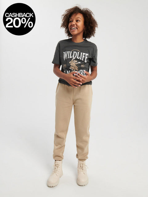 Person wearing a dark gray t-shirt with text and graphics, beige pants, and white shoes on a light gray background