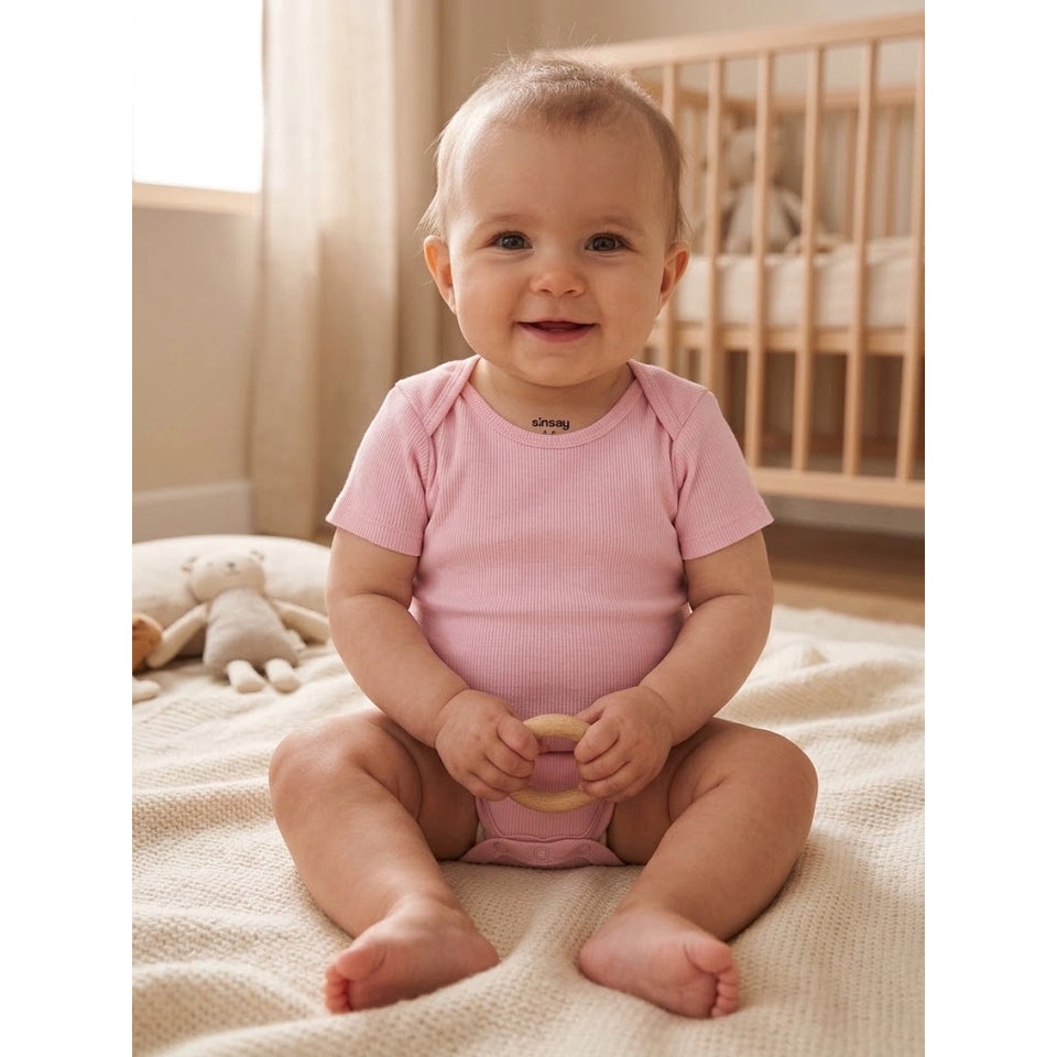 A baby sitting on a cream textured blanket in a nursery, wearing a light pink short-sleeve ribbed bodysuit and holding a wooden teething ring.
