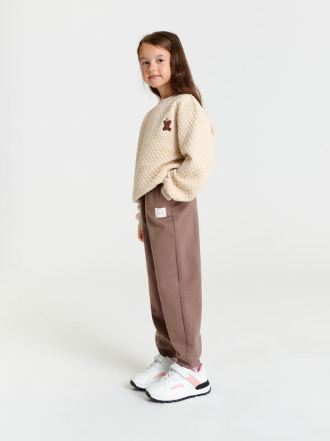 Young girl wearing a beige sweater and brown pants on a white background