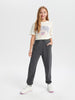 Young girl wearing a white t-shirt with text and graphics, dark gray pants, and white sneakers on a white background