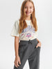 Young girl wearing a white t-shirt with colorful graphics and gray pants on a light gray background