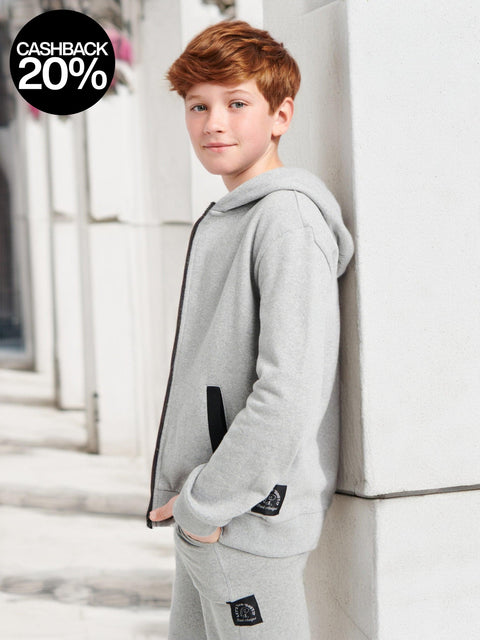 Young boy wearing a gray hoodie and pants leaning against a white wall.