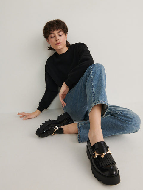 Person wearing black loafers with gold accents and blue jeans sitting on a white floor.