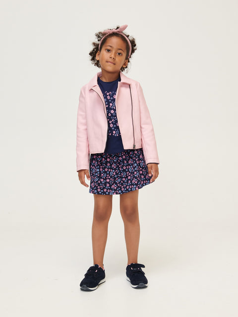 Young girl wearing a pink jacket, navy floral dress, and skirt on a white background