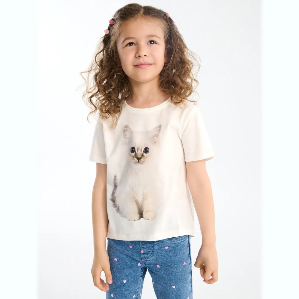A young girl with curly brown hair wearing a cream-colored t-shirt with a large printed kitten graphic and blue jeans with small pink hearts, standing against a plain white background.
