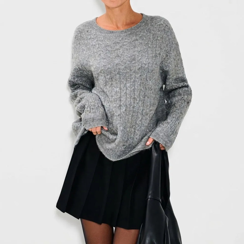 A woman wearing an oversized heather gray knit sweater and a black pleated skirt, holding a black bag against a plain light gray studio background.