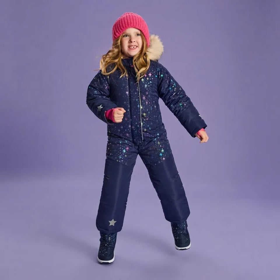 A young girl wearing a dark navy blue snowsuit with a starry pattern, a pink knit beanie, and dark winter boots, posing against a solid purple studio background.