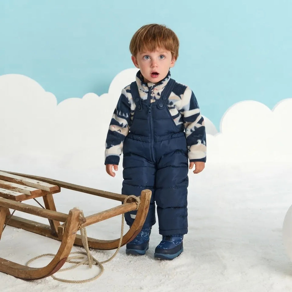 A toddler stands in a snowy studio setting wearing a navy blue puffer snowsuit over a navy and cream patterned long-sleeve top, with blue boots. A wooden sled is to the left, and the background features a light blue sky and white cloud cutouts.