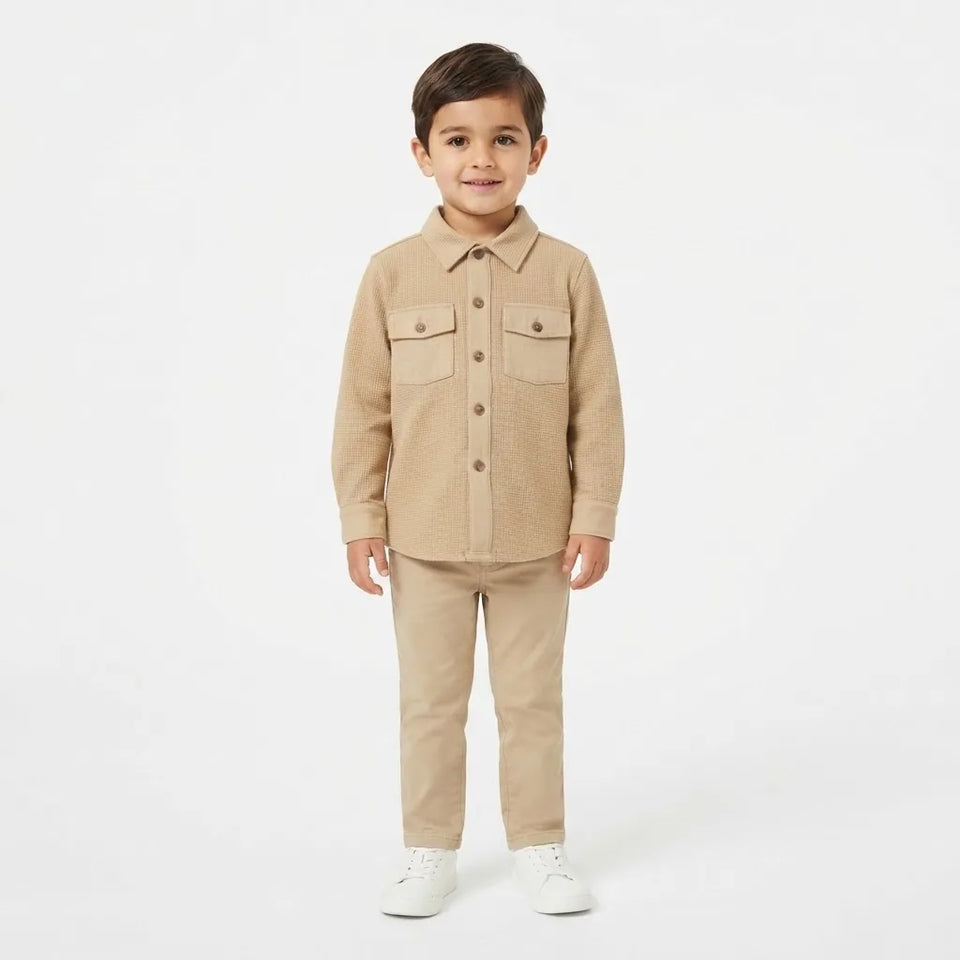 A young boy modeling a beige two-piece outfit consisting of a long-sleeve button-up shirt and matching trousers, paired with white sneakers, against a plain white studio background.