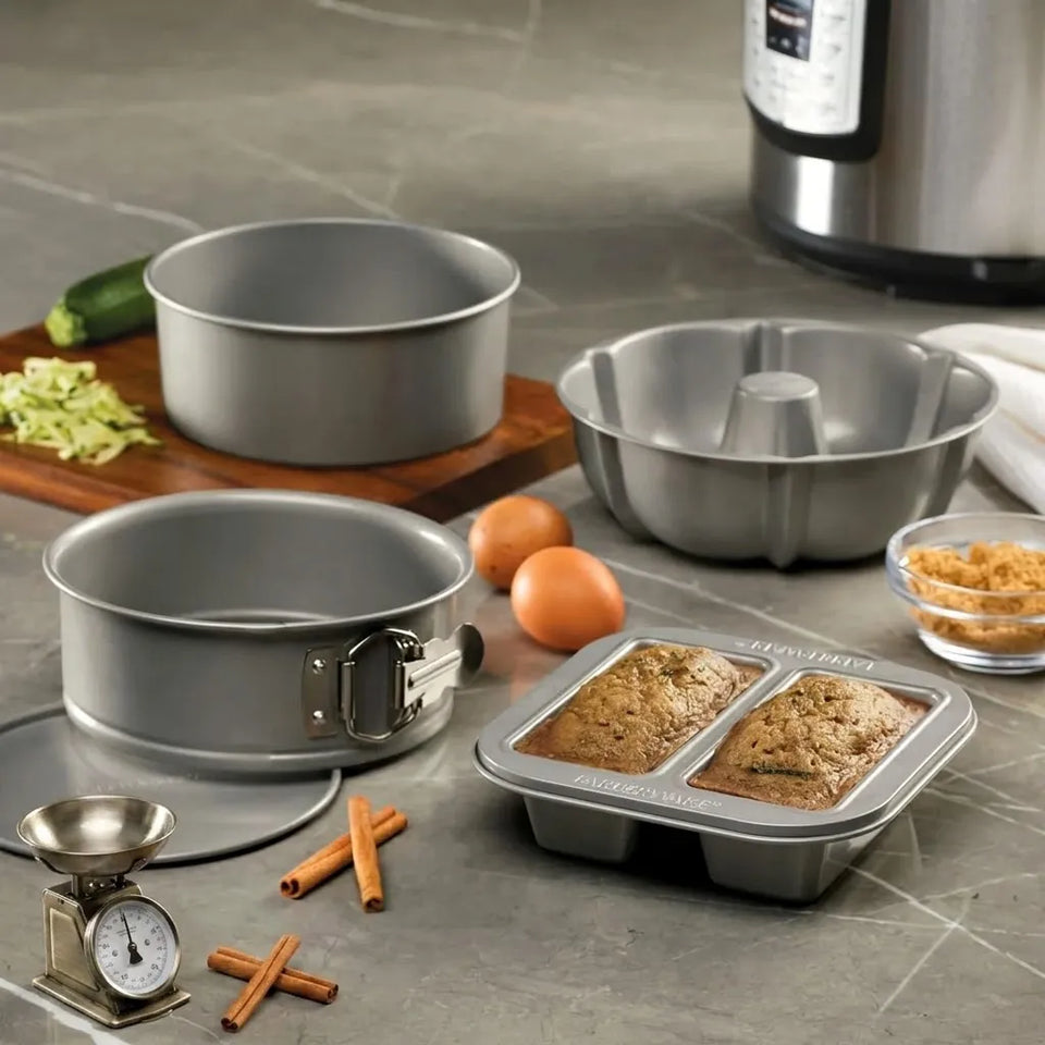 A set of stainless steel baking pans and a loaf pan shown on a gray countertop with baking ingredients and a partially baked double loaf inside. The set includes two round springform pans, a bundt pan, and a rectangular loaf pan with baked bread.