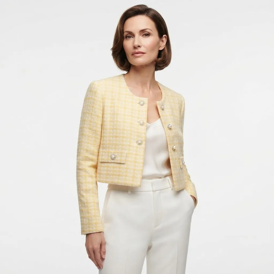 A woman modeling a cropped yellow and white check tweed jacket with gold-tone buttons, worn over a white top and white trousers, against a plain white studio background.