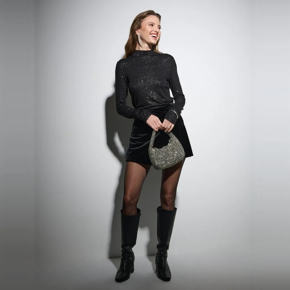 A woman stands against a light gray studio wall wearing a black long-sleeve sequined top, a black mini skirt, black knee-high boots, and holding a silver glitter handbag.