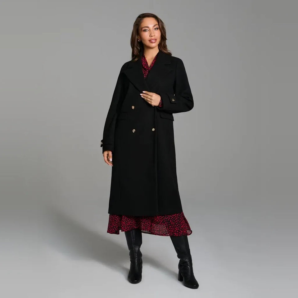 A woman modeling a long black double-breasted coat over a red and black patterned dress, paired with black ankle boots, against a solid gray studio background.