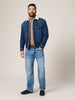Man wearing a blue denim jacket with a white collar, beige sweater, and blue jeans on a plain background