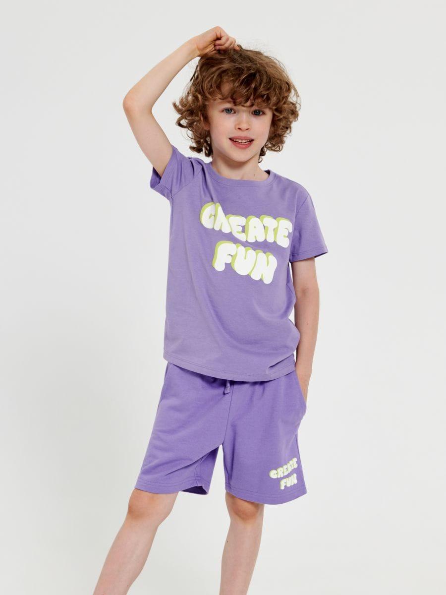 A boy wearing a purple short-sleeve t-shirt with text on it and matching purple shorts.