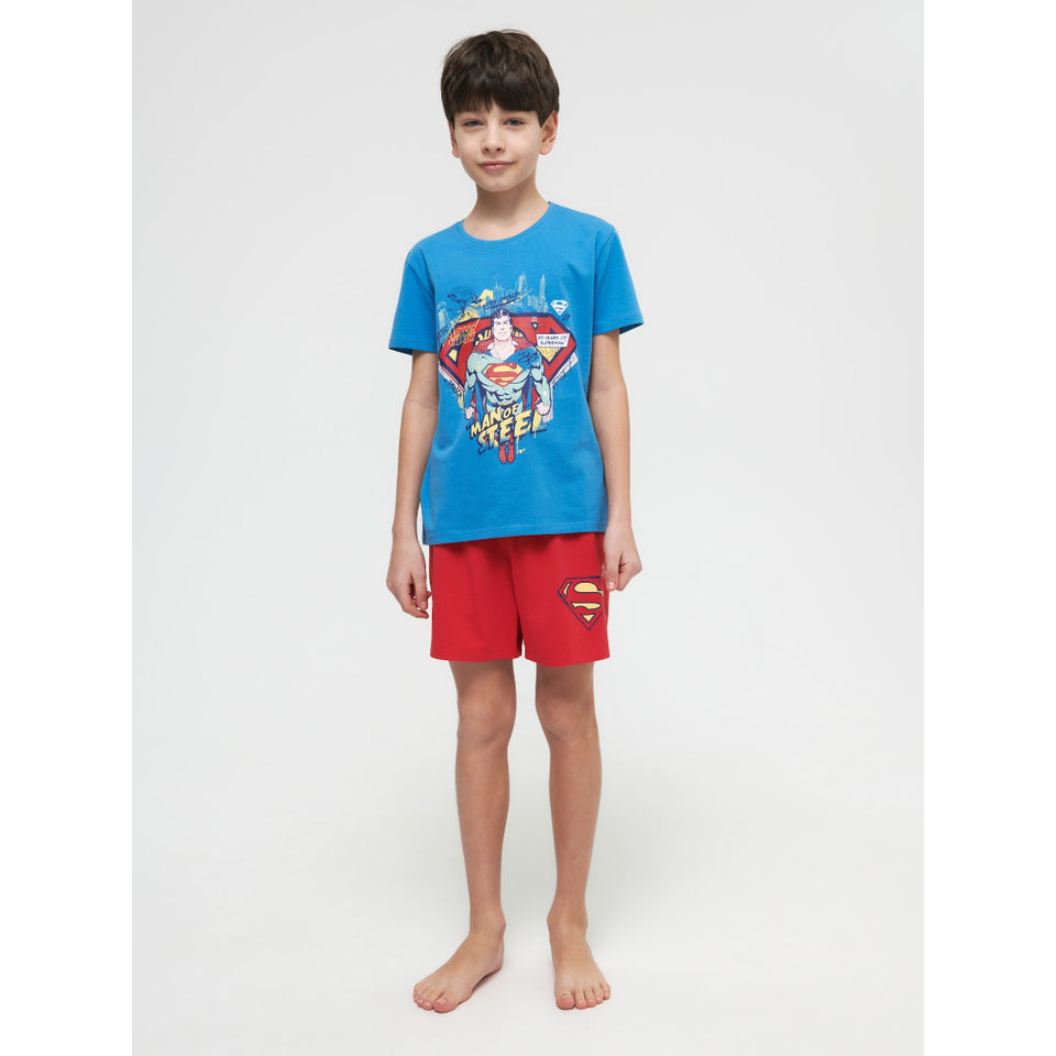 A young boy stands barefoot against a light gray studio background, wearing a bright blue Superman graphic T-shirt and red shorts with a Superman logo patch.