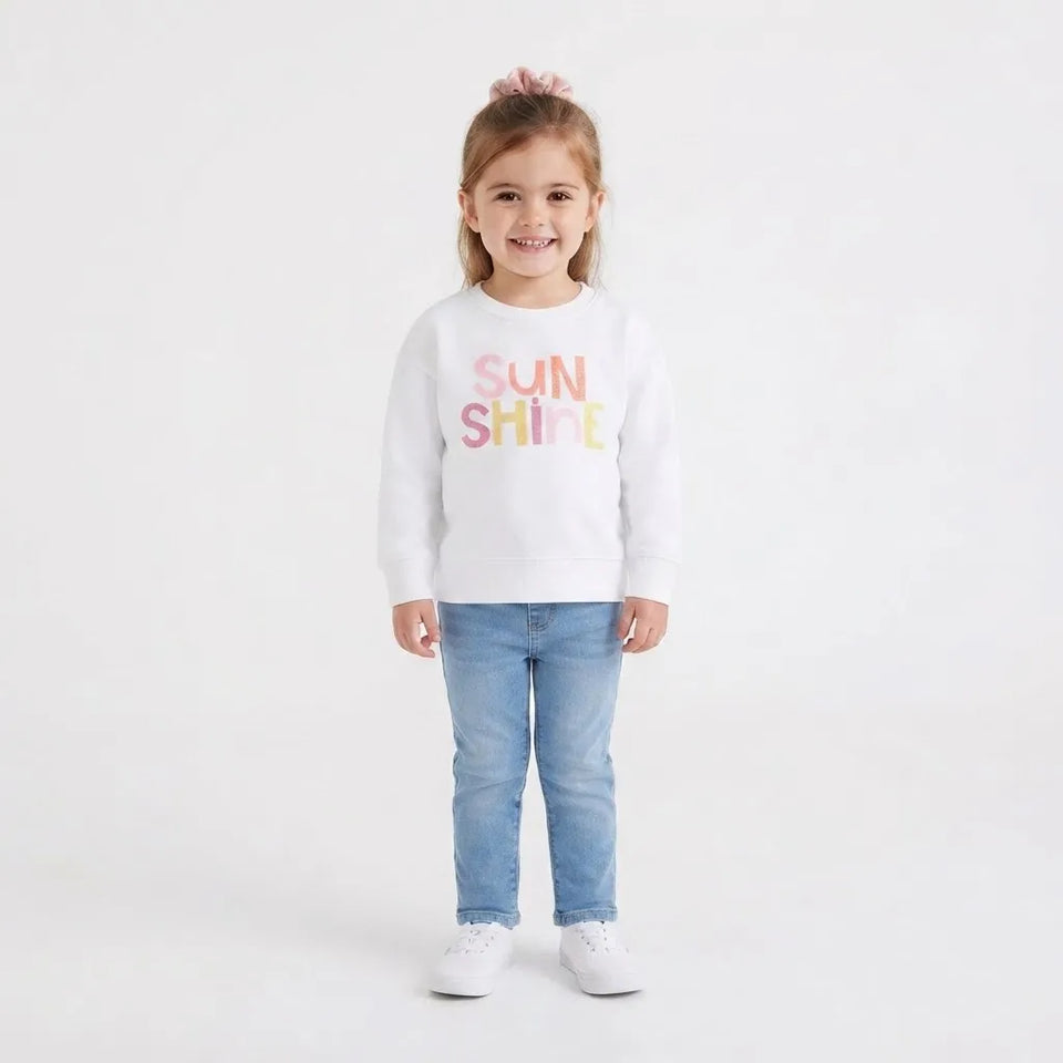 A young girl with a pink hair bow stands against a white studio background wearing a white crewneck sweatshirt with 'SUN SHINE' printed in pastel pink, orange, and yellow letters, paired with light blue jeans and white sneakers.