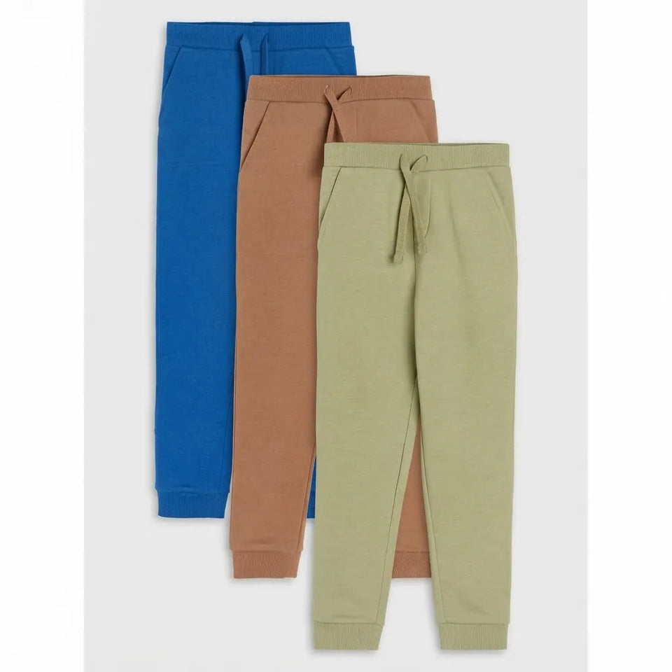 Three pairs of children's jogger sweatpants in blue, tan, and sage green, arranged in a vertical overlapping stack on a light gray background.