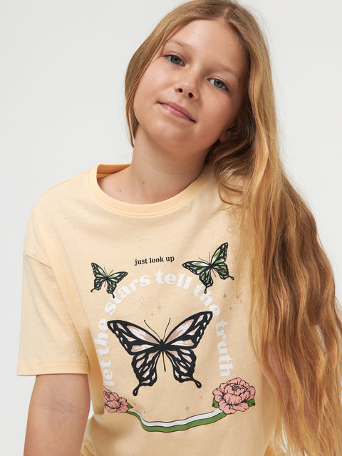 Young girl wearing a light yellow t-shirt with butterfly designs and text.