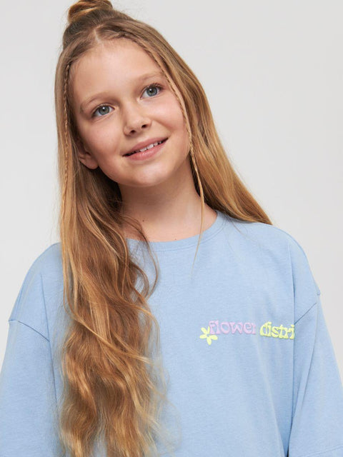 Young girl wearing a light blue sweatshirt with a logo on a plain background