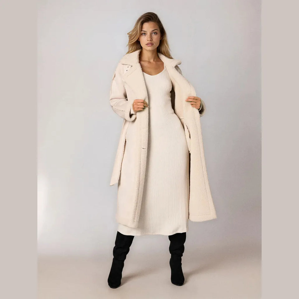 A woman models a long cream-colored textured coat over a matching ribbed knit dress, paired with black knee-high boots, against a neutral studio background.