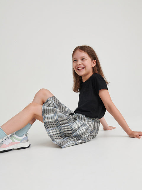 Young girl sitting on a white floor wearing a black t-shirt and plaid skirt.