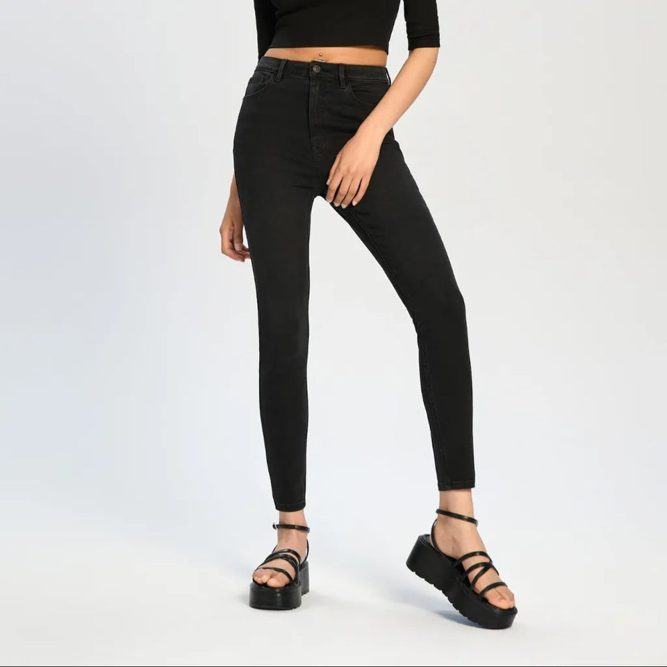 A model wearing black skinny jeans, a black cropped long-sleeve top, and black strappy platform sandals, standing against a light gray studio background.