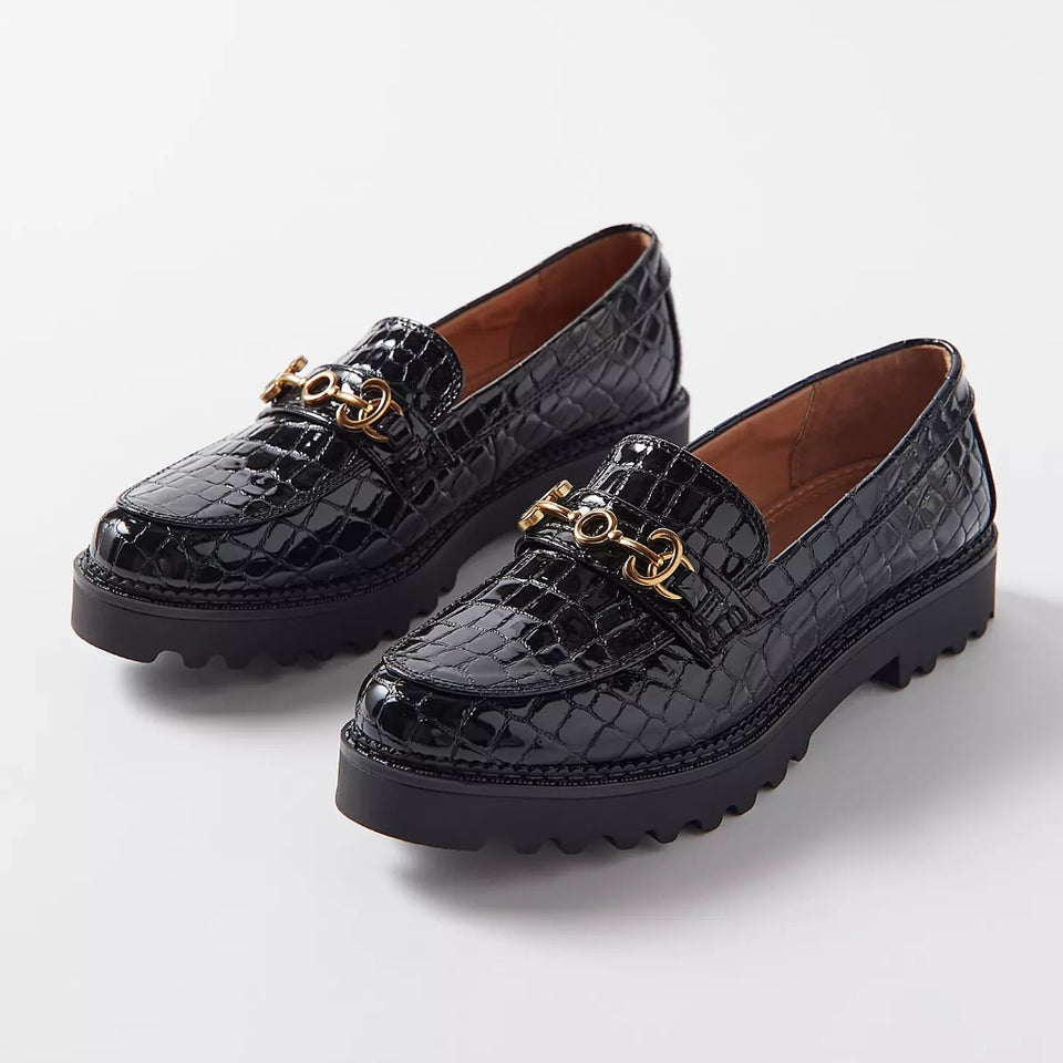 A pair of black crocodile-embossed leather loafers with a gold-tone chain detail across the vamp and chunky lug soles, photographed on a light gray studio background.