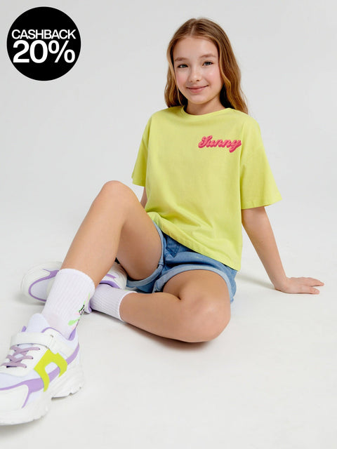 Young girl wearing a yellow t-shirt with text, denim shorts, and colorful sneakers on a white background