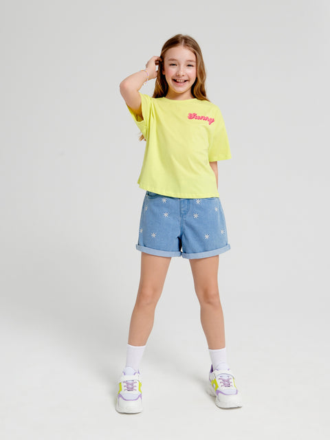Young girl wearing a yellow t-shirt and blue denim shorts with white patterns on a white background