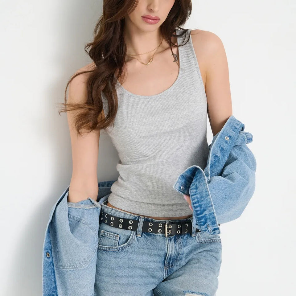 A woman wearing a light gray ribbed tank top, light-wash blue jeans, a black studded belt, and holding a light blue denim shirt over her shoulder, posed against a plain white wall.