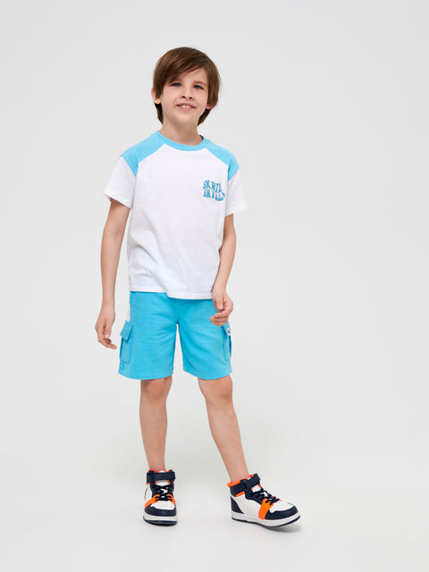 Child wearing a white and blue shirt with blue shorts on a light gray background