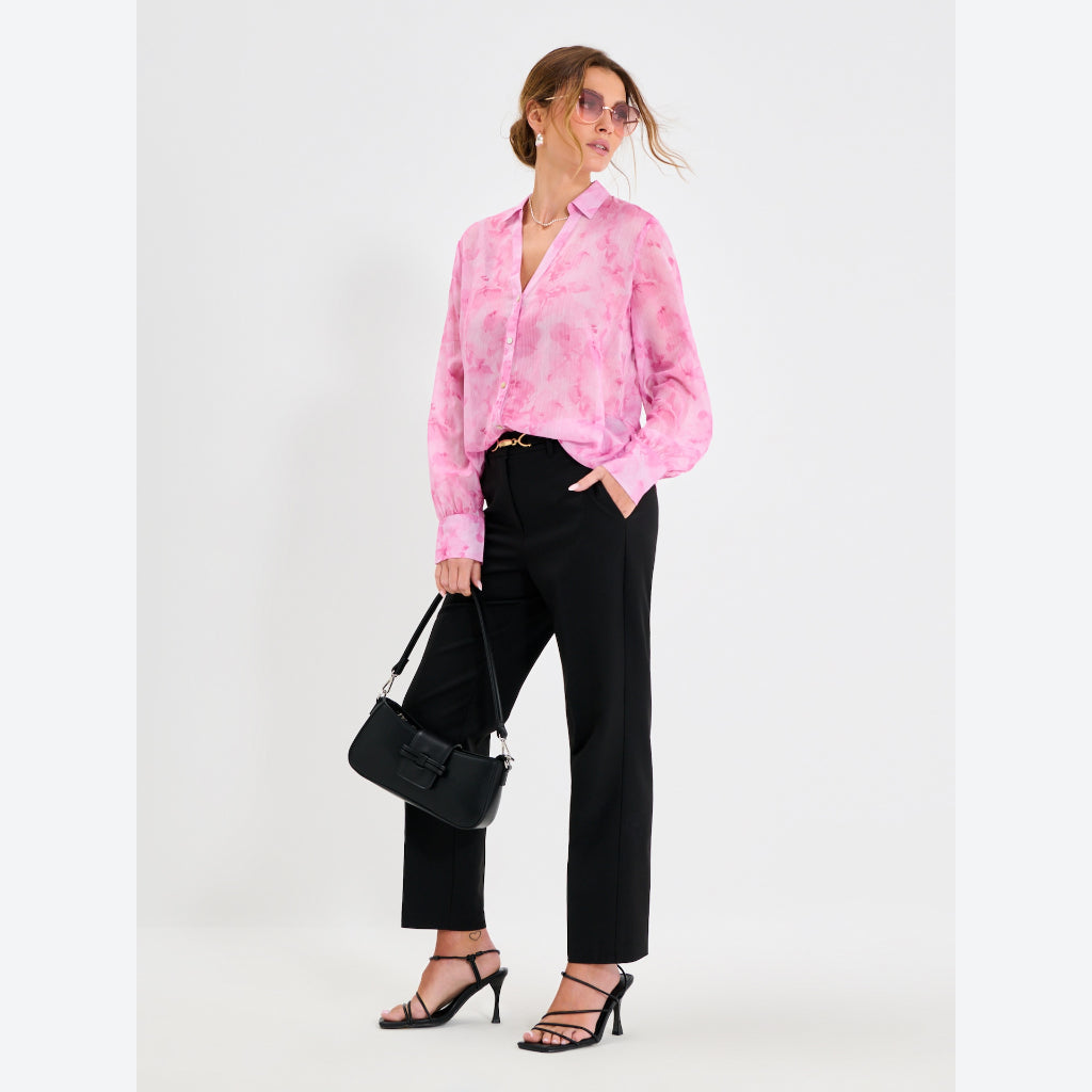 A woman modeling a pink and white tie-dye long-sleeve button-up blouse, paired with black cropped trousers, black strappy heels, and carrying a black handbag, set against a plain light gray studio background.