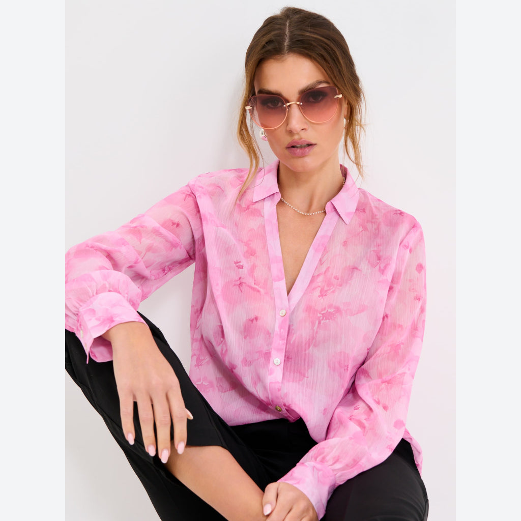 A woman wearing a pink long-sleeve button-up blouse with a subtle tonal floral pattern, paired with black pants and gold-framed sunglasses, posed against a light gray studio background.