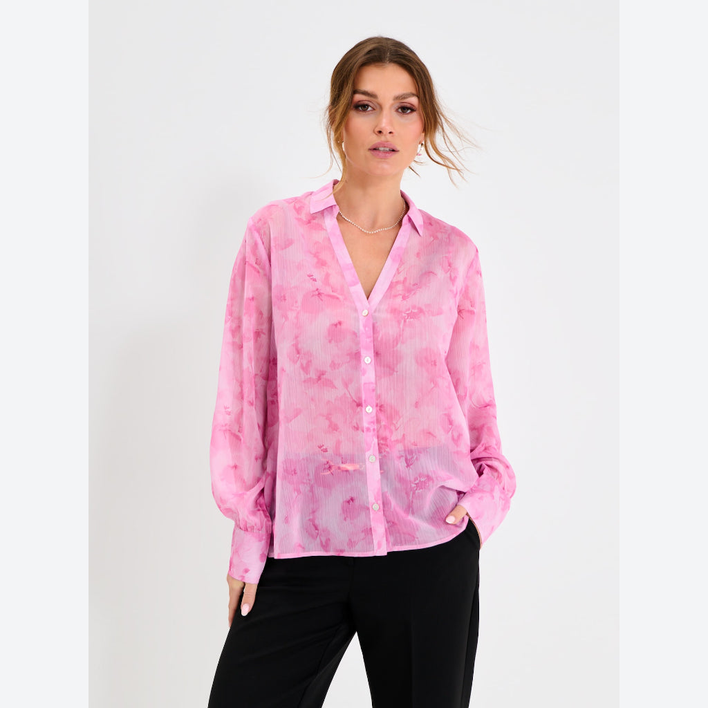 A woman modeling a pink and light pink watercolor-patterned long-sleeve button-up shirt with black pants, set against a plain light gray background.