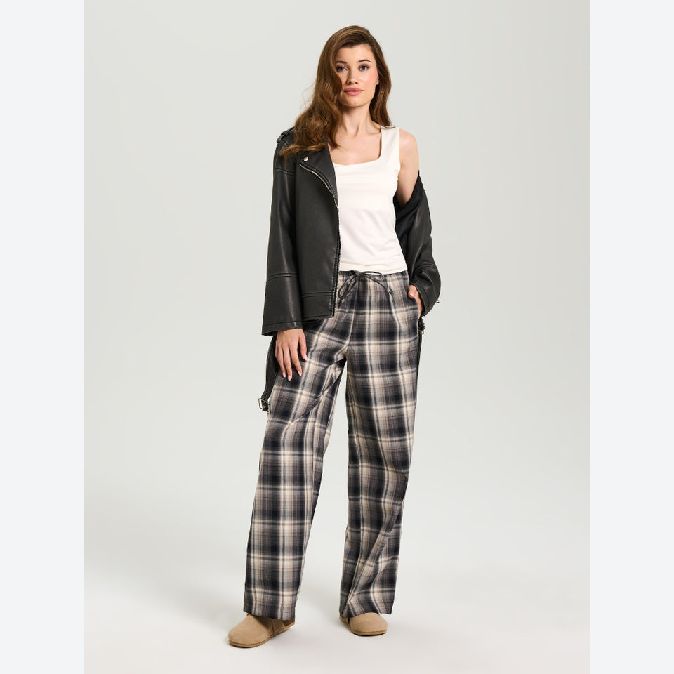 A model stands in a studio wearing a black leather-look moto jacket over a white tank top, paired with wide-leg plaid trousers in gray, black, and beige, and tan shoes.