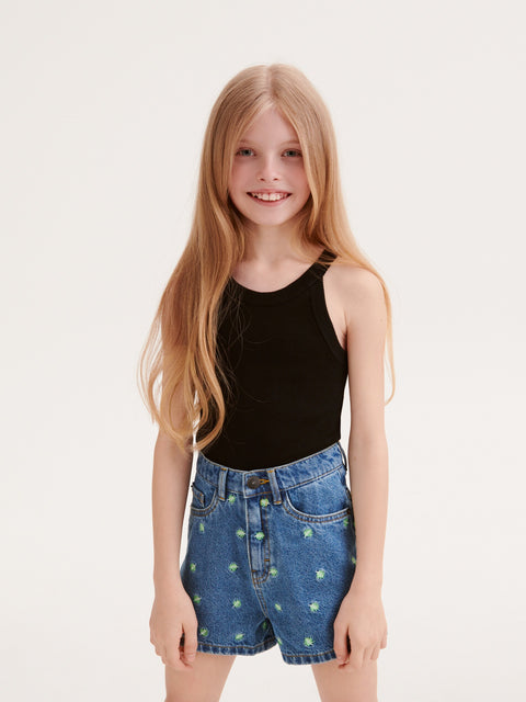 Young girl wearing a black tank top and denim shorts with green patterns on a white background
