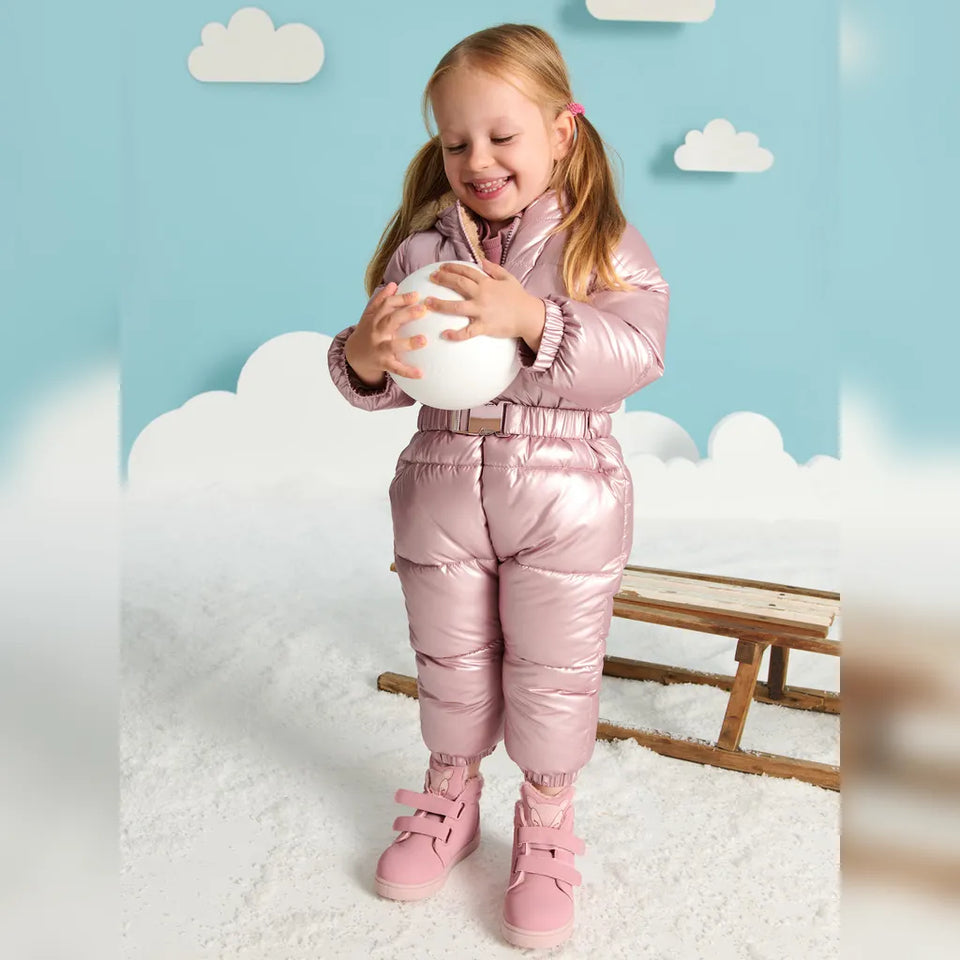 A young girl stands in a studio setting wearing a shiny metallic pink puffer snowsuit with matching pink boots, holding a white ball. The background features a light blue sky backdrop with white cloud cutouts, a wooden bench, and a white textured floor.