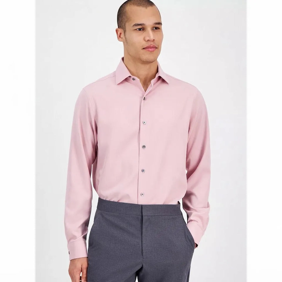 A man wearing a light pink long-sleeve button-down dress shirt and dark gray trousers, standing against a plain light gray studio background.