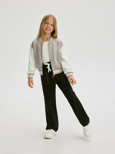 Young girl wearing a gray and white varsity jacket, black pants, and white sneakers on a white background