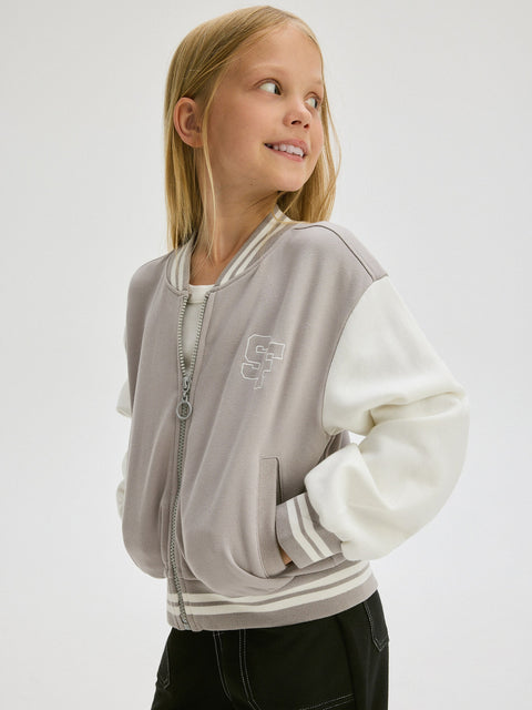 Young girl wearing a gray and white varsity jacket on a light gray background