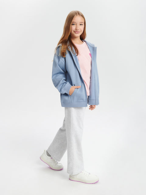 Young girl wearing a light blue jacket over a pink shirt, white pants, and white sneakers on a white background