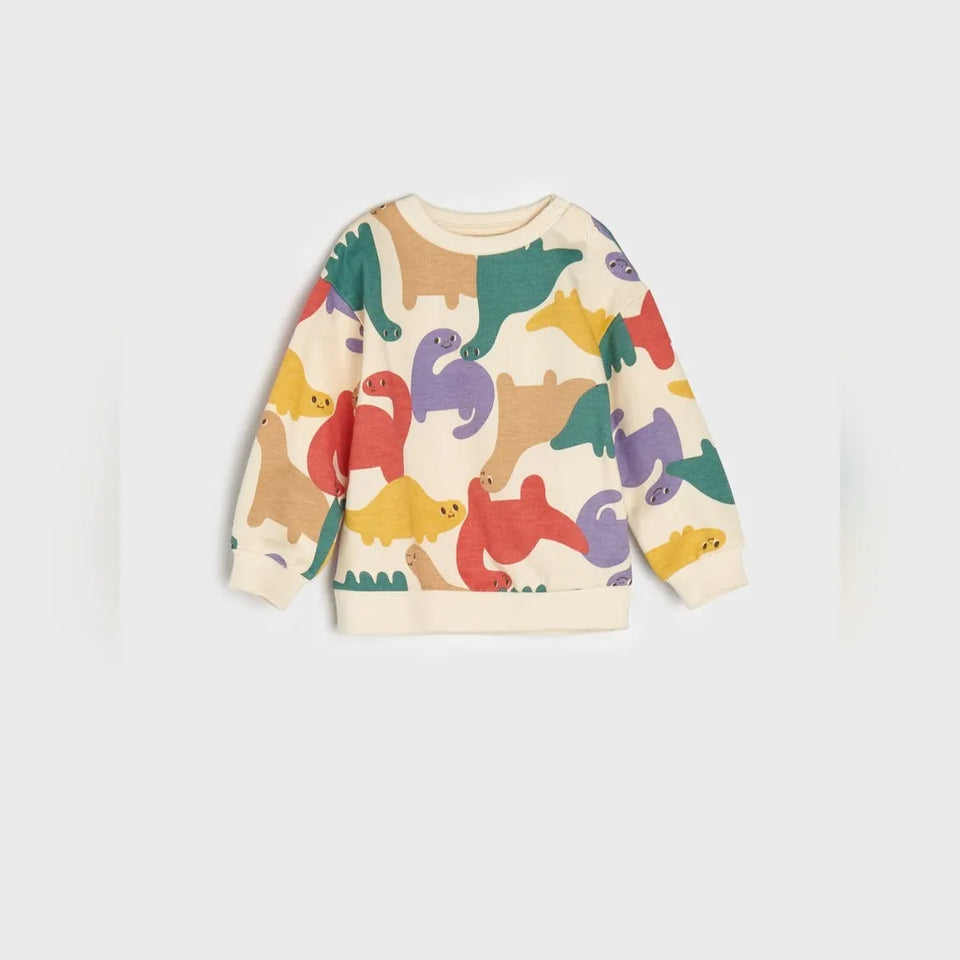 A cream-colored long-sleeve sweatshirt featuring a colorful dinosaur pattern in green, purple, red, yellow, and brown, displayed against a light gray background.