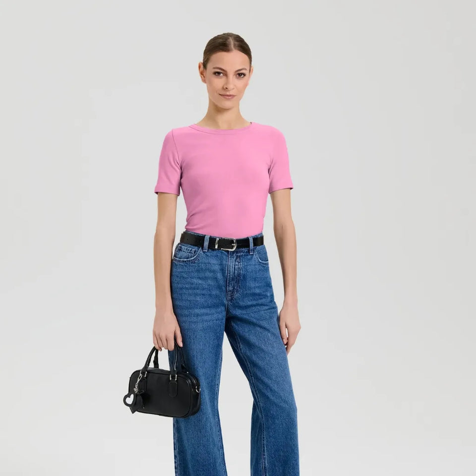 A woman modeling a short-sleeve pink crewneck t-shirt, paired with high-waisted blue jeans, a black belt, and holding a small black handbag against a light gray studio background.