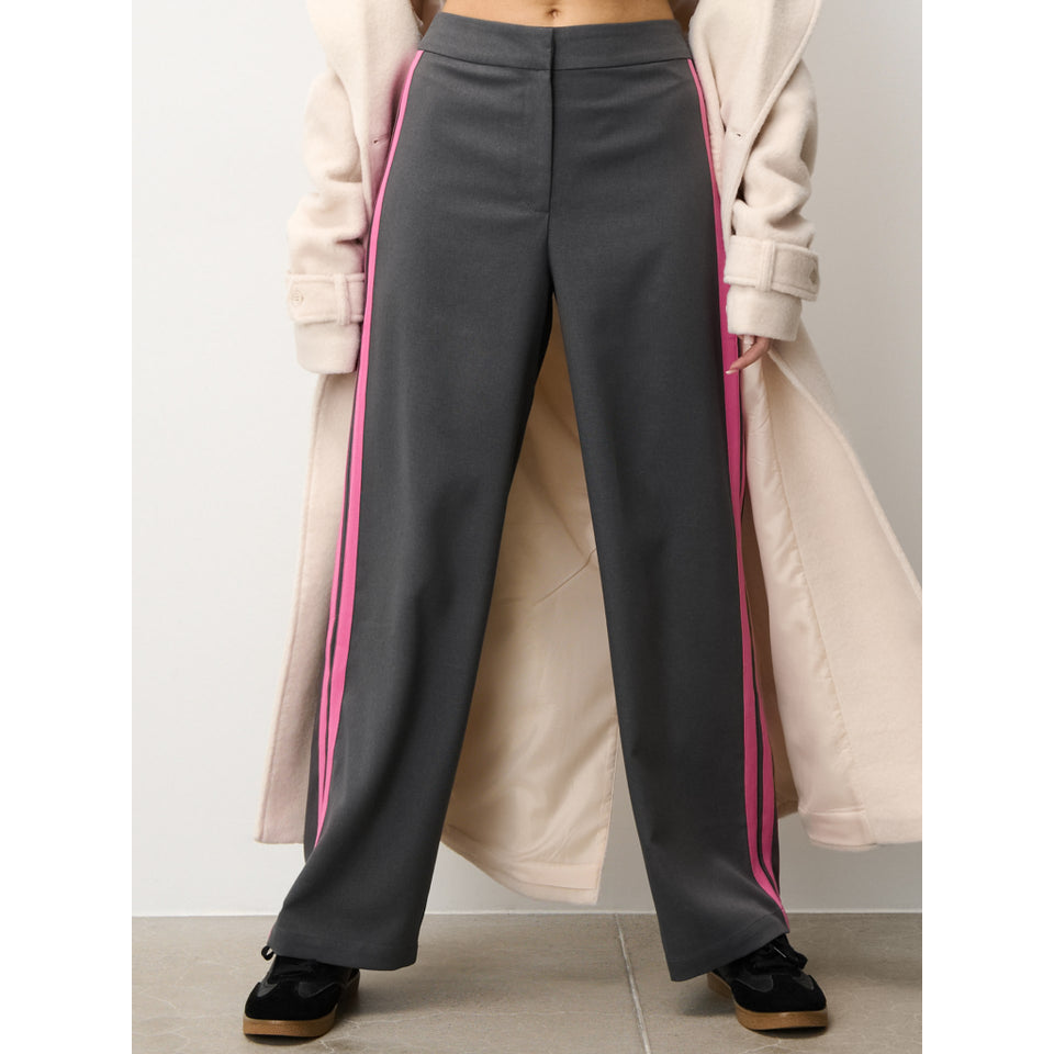 A model is wearing dark gray wide-leg trousers with vibrant pink side stripes, styled with a beige trench coat and black shoes.