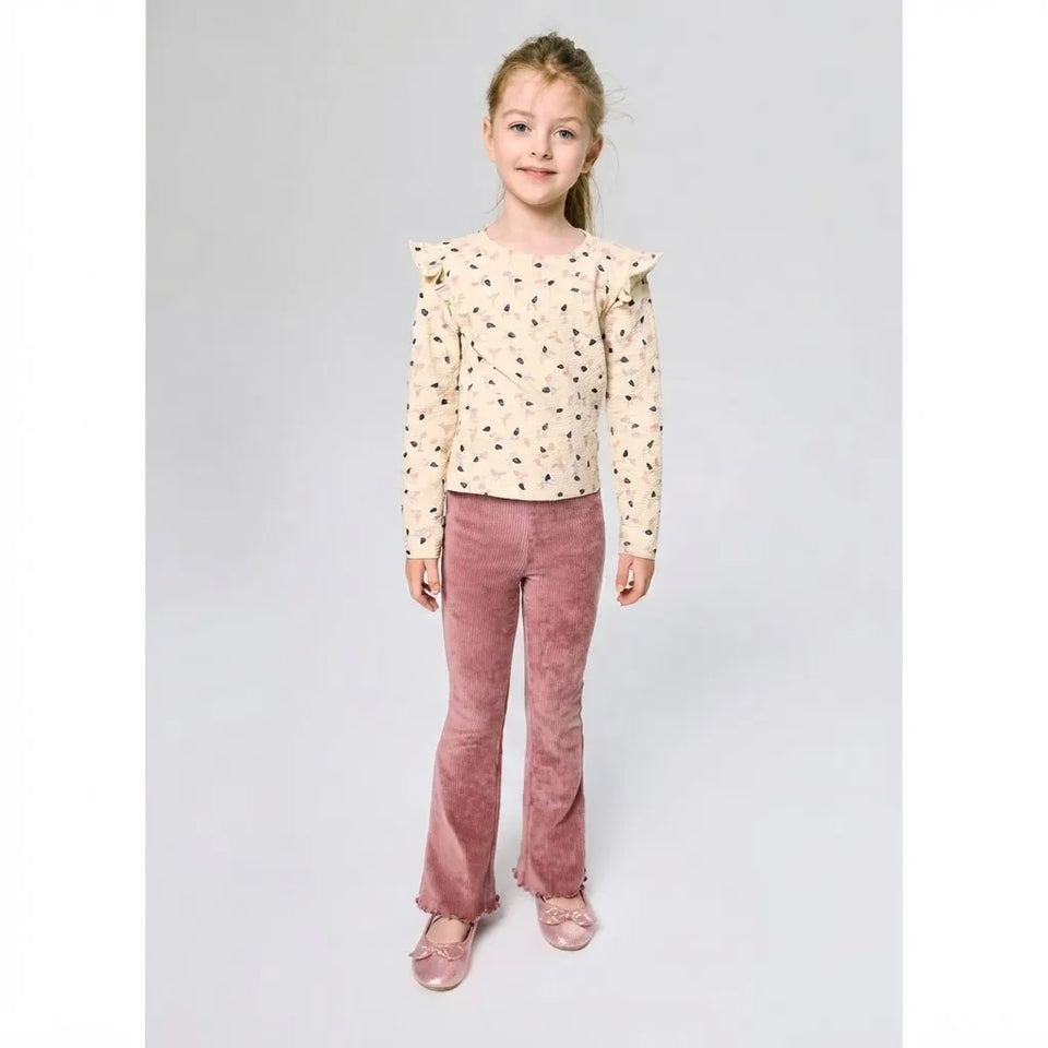 A young girl stands against a light gray studio background wearing a cream long-sleeve blouse with small black motifs and ruffled shoulders, paired with dusty rose corduroy pants and matching pink shoes.