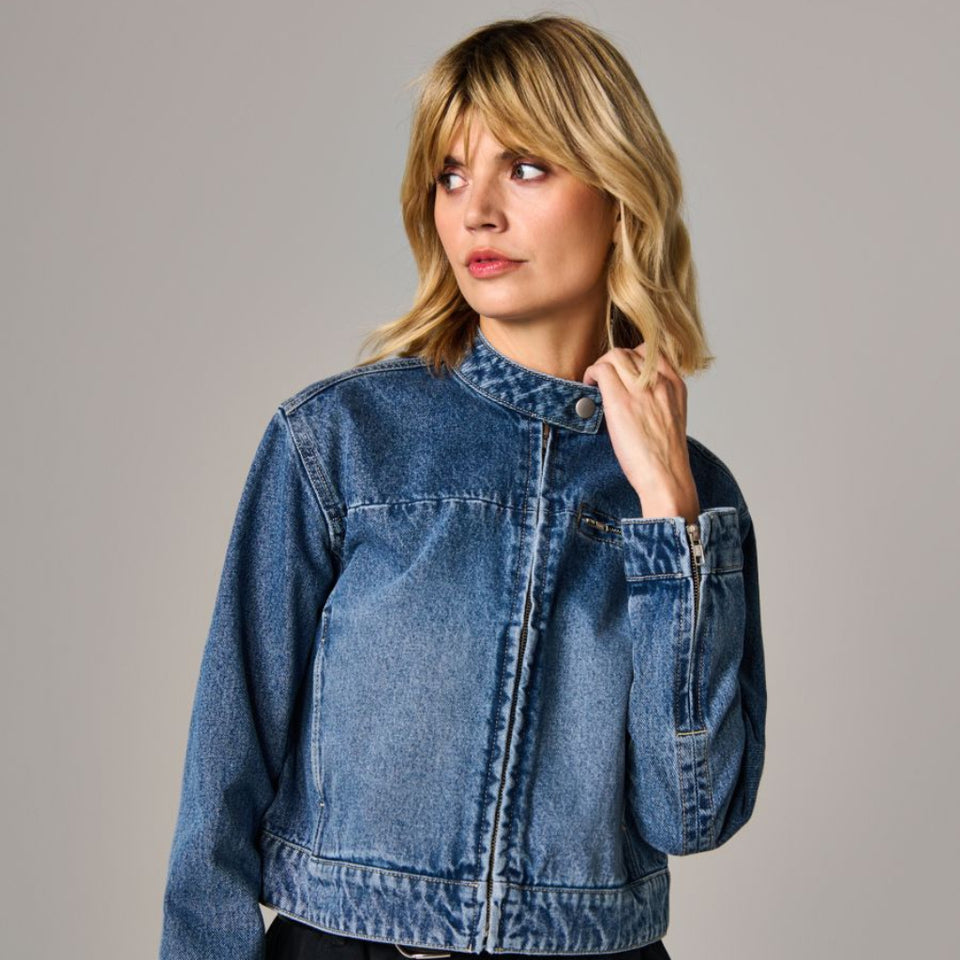 A woman wearing a cropped blue denim jacket with a stand collar and front button closure, paired with black pants, photographed against a plain light gray studio background.
