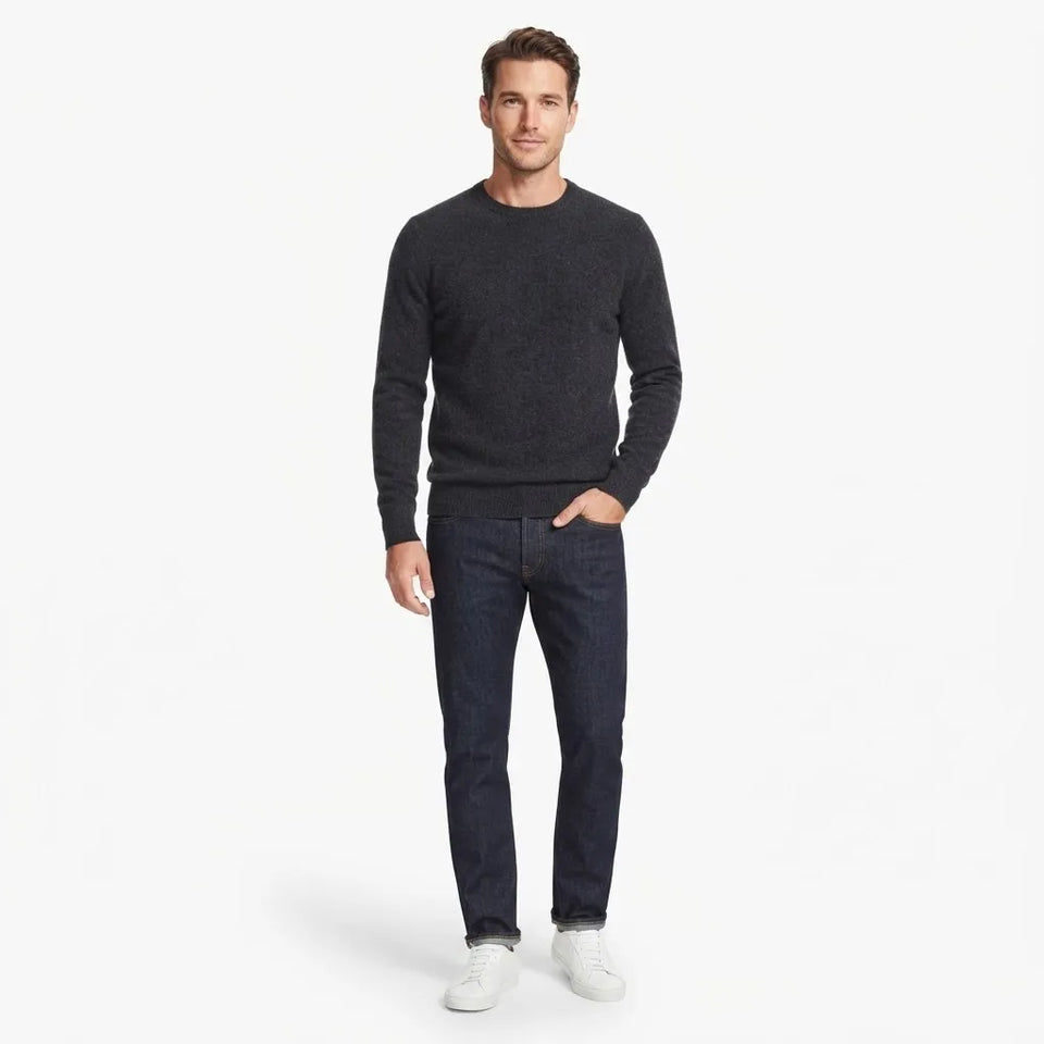 A man modeling a dark charcoal gray crewneck sweater, dark blue jeans, and white sneakers against a plain white studio background.