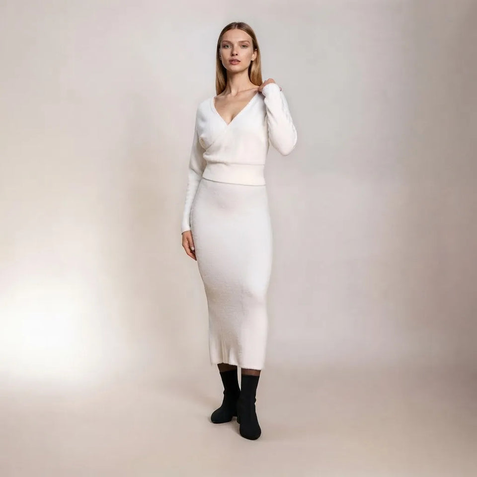 A woman modeling a white ribbed knit long-sleeve midi dress with a wrap-style V-neck bodice, paired with black ankle boots, against a plain light beige studio backdrop.