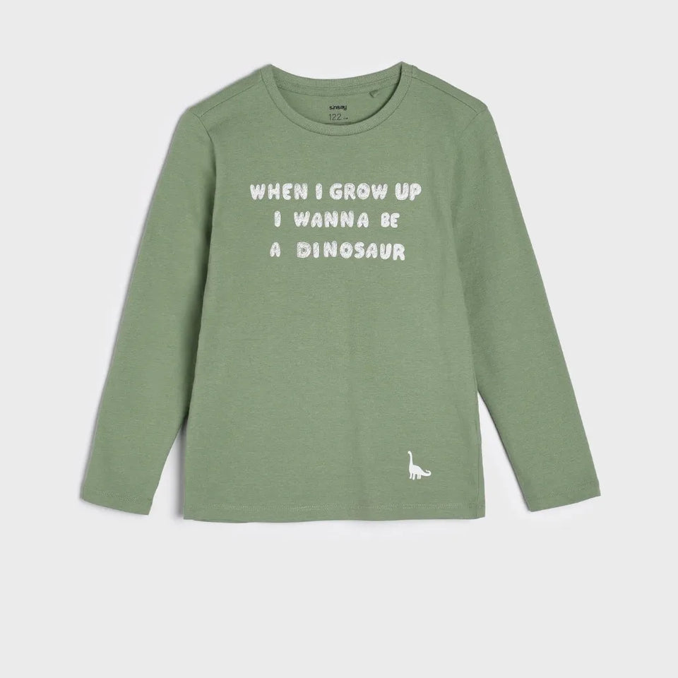 A sage green long-sleeve crewneck shirt for kids, featuring the white text 'WHEN I GROW UP I WANNA BE A DINOSAUR' across the chest and a small white dinosaur silhouette on the lower left hem.